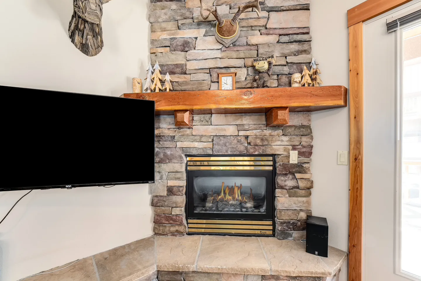 Corner stone fireplace with a thick wood mantel, lit gas flames, and a mounted flat-screen TV to the left.