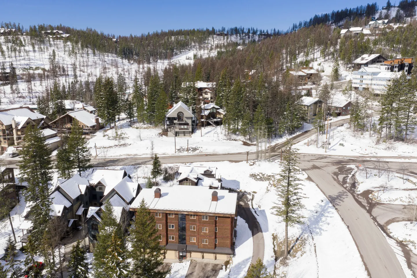 Aerial view of a multi-story building and neighboring mountain lodges surrounded by snow-covered ground and evergreen trees.