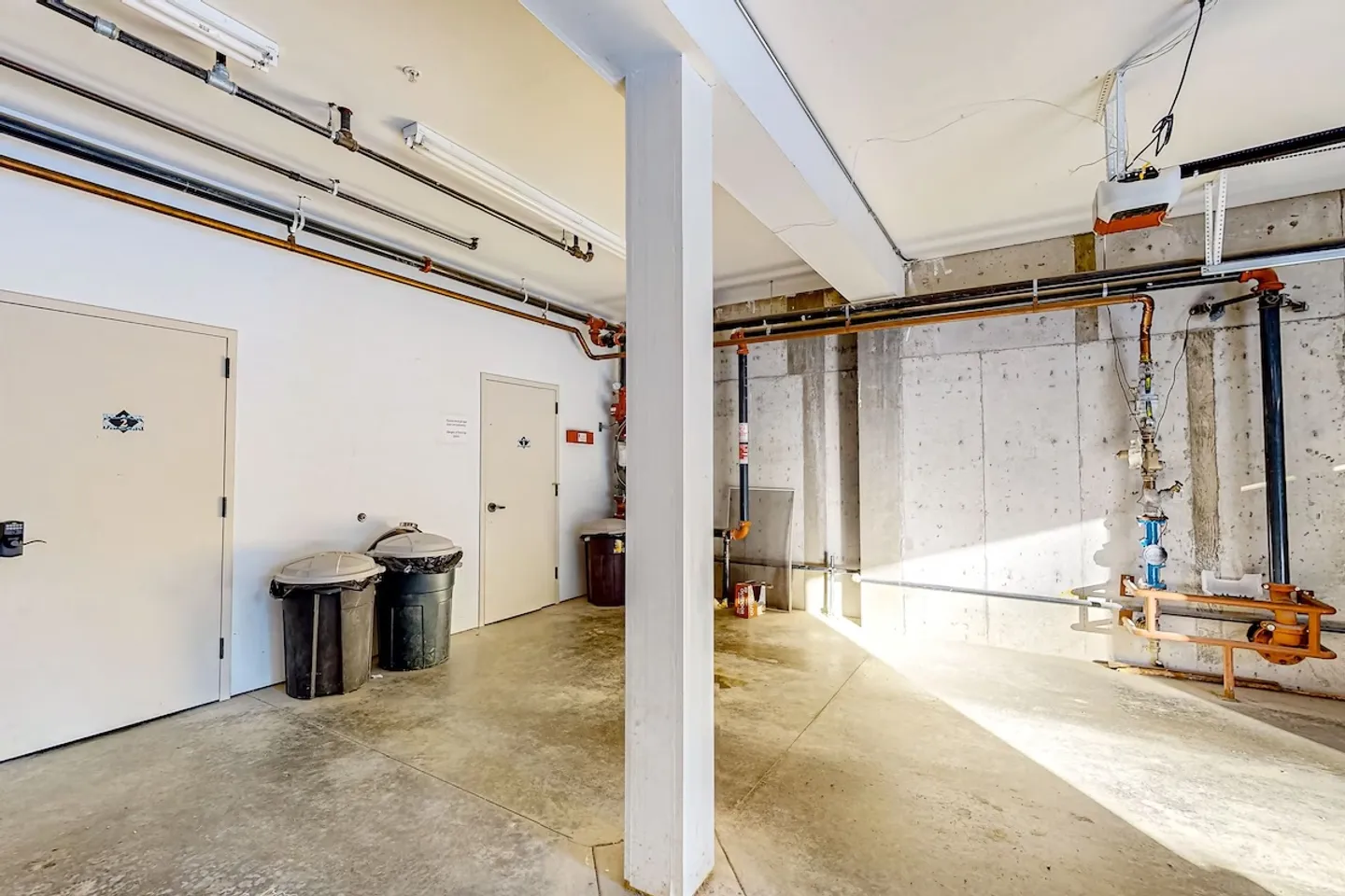 Utility area or garage interior featuring concrete floors, exposed pipes, support pillars, trash bins, and closed doors.