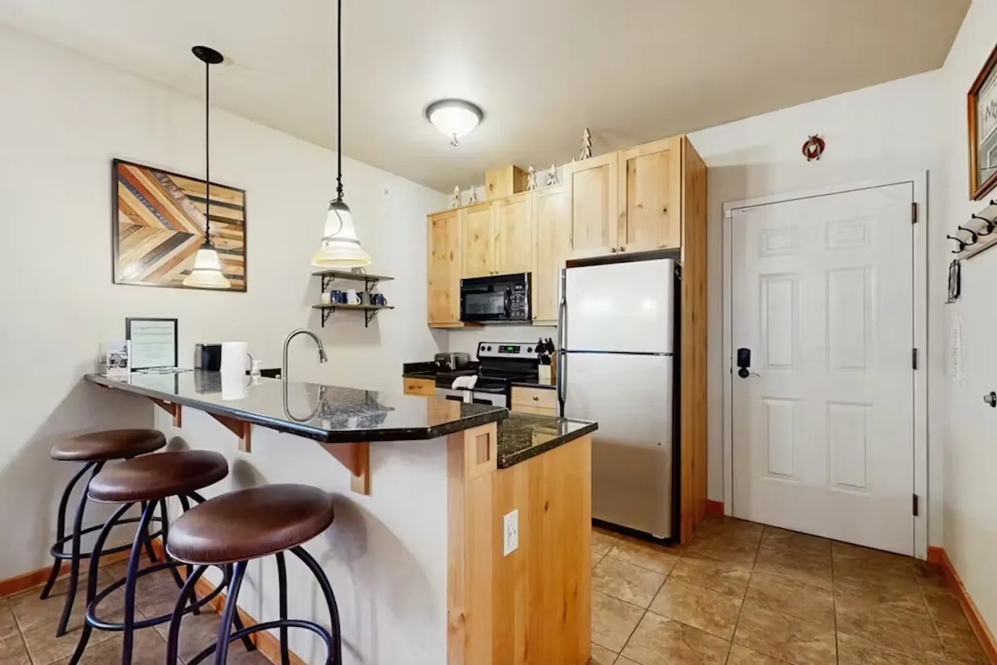 Kitchen with light wood cabinets, dark countertops, a breakfast bar with three stools, and stainless steel appliances.