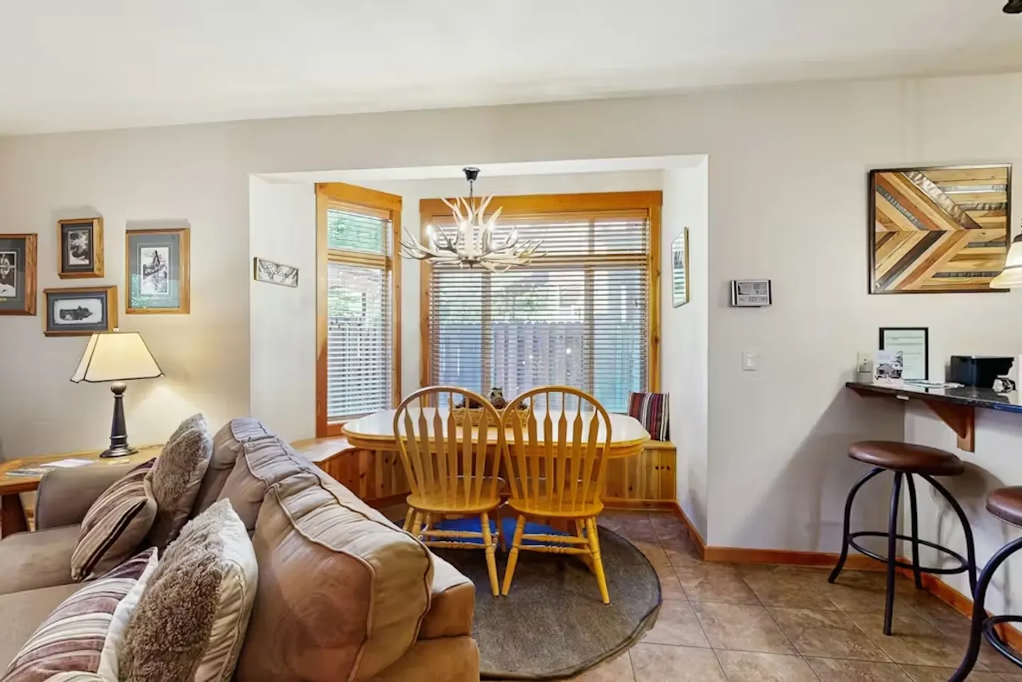 Dining nook with a wooden table, chairs, and built-in bench seating beside large windows, viewed from the adjacent living area.