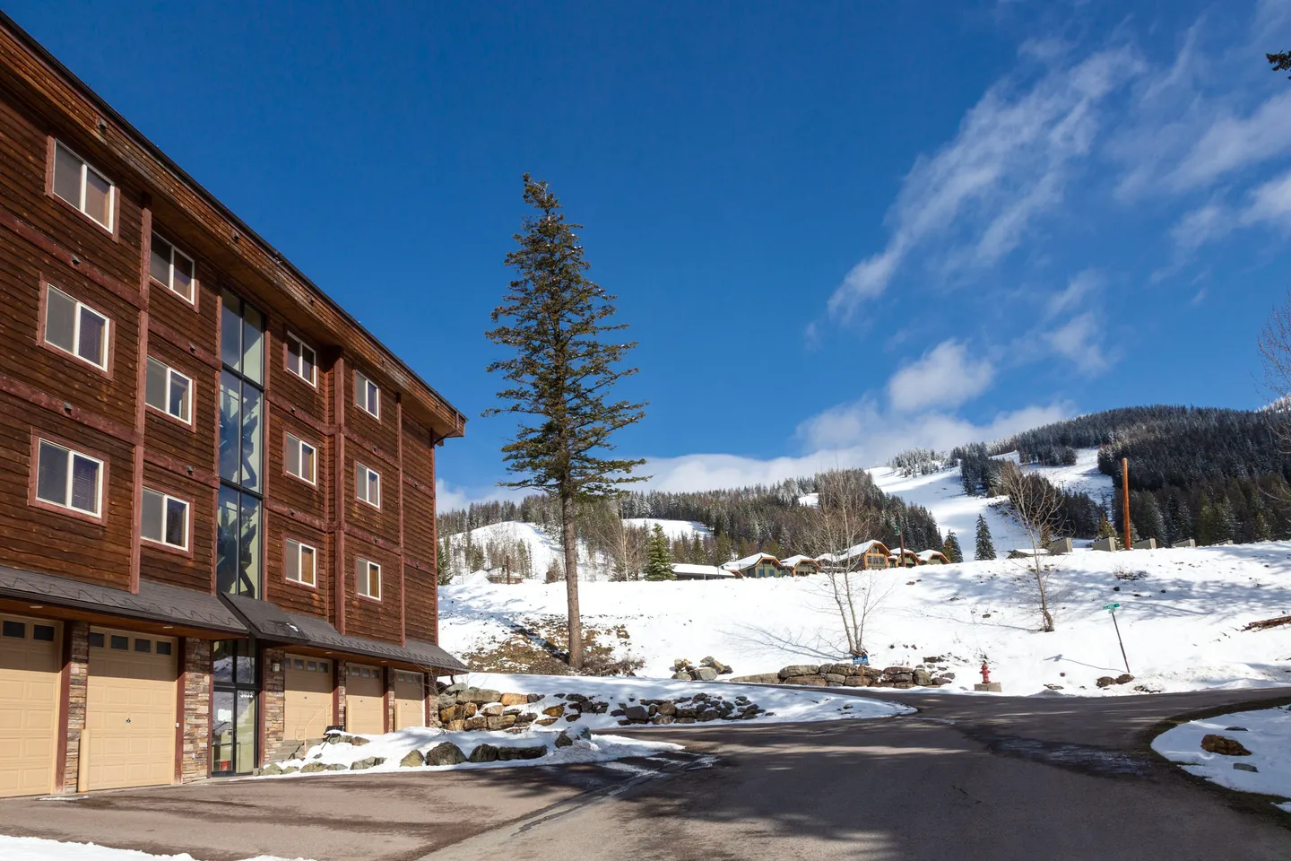 Exterior of a multi-story wood-sided building with garage doors, adjacent to a paved driveway and snow-covered hills under a blue sky.