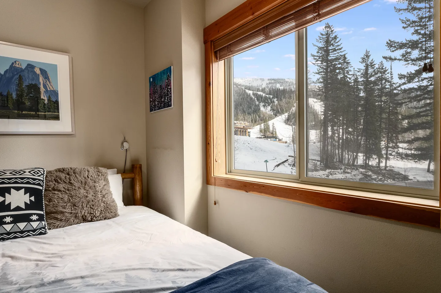 Bedroom with a wooden log frame bed next to a large window overlooking snow-covered ski slopes and pine trees.