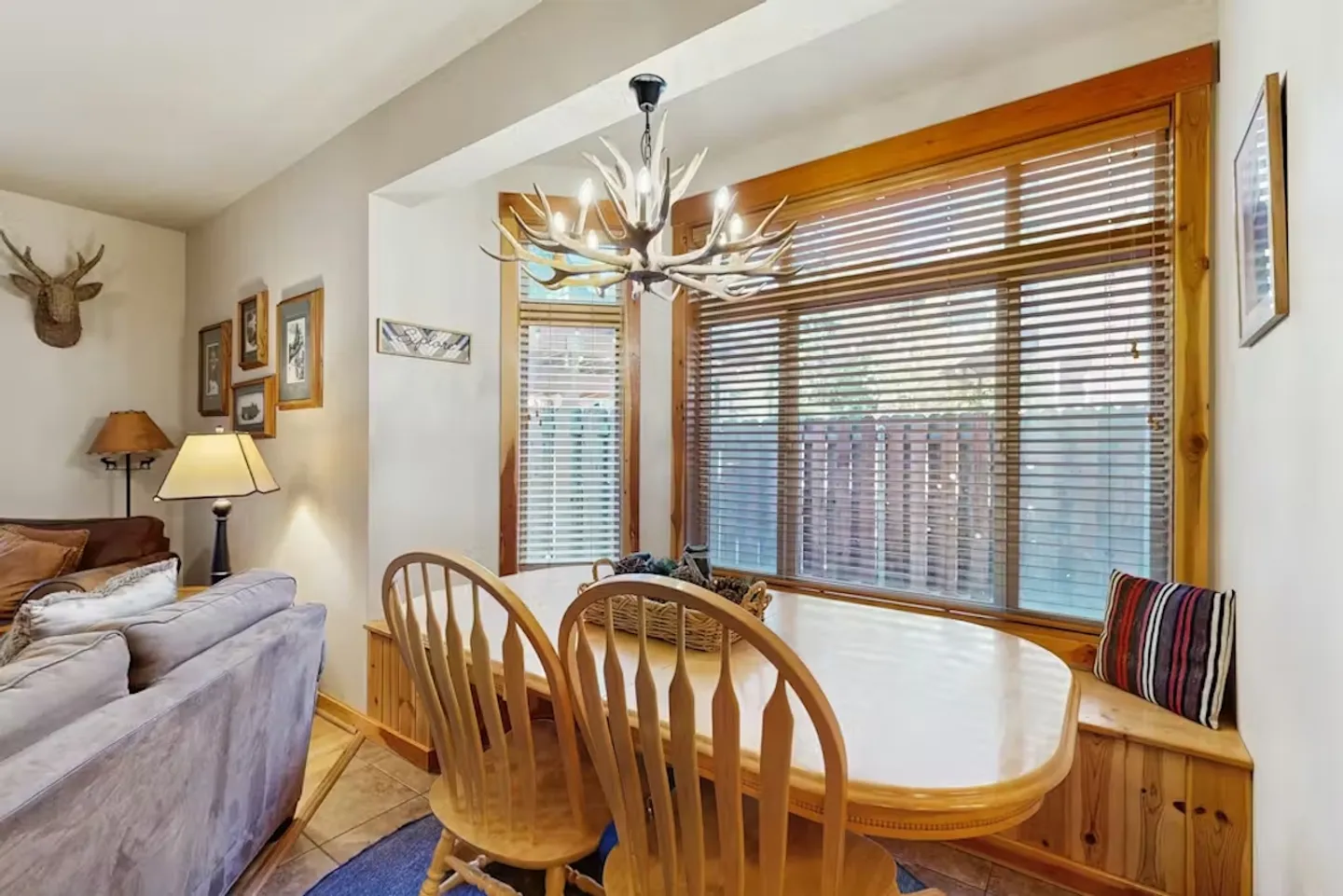 Dining nook with a wooden table, built-in window bench, wooden chairs, and an antler chandelier next to a living area.
