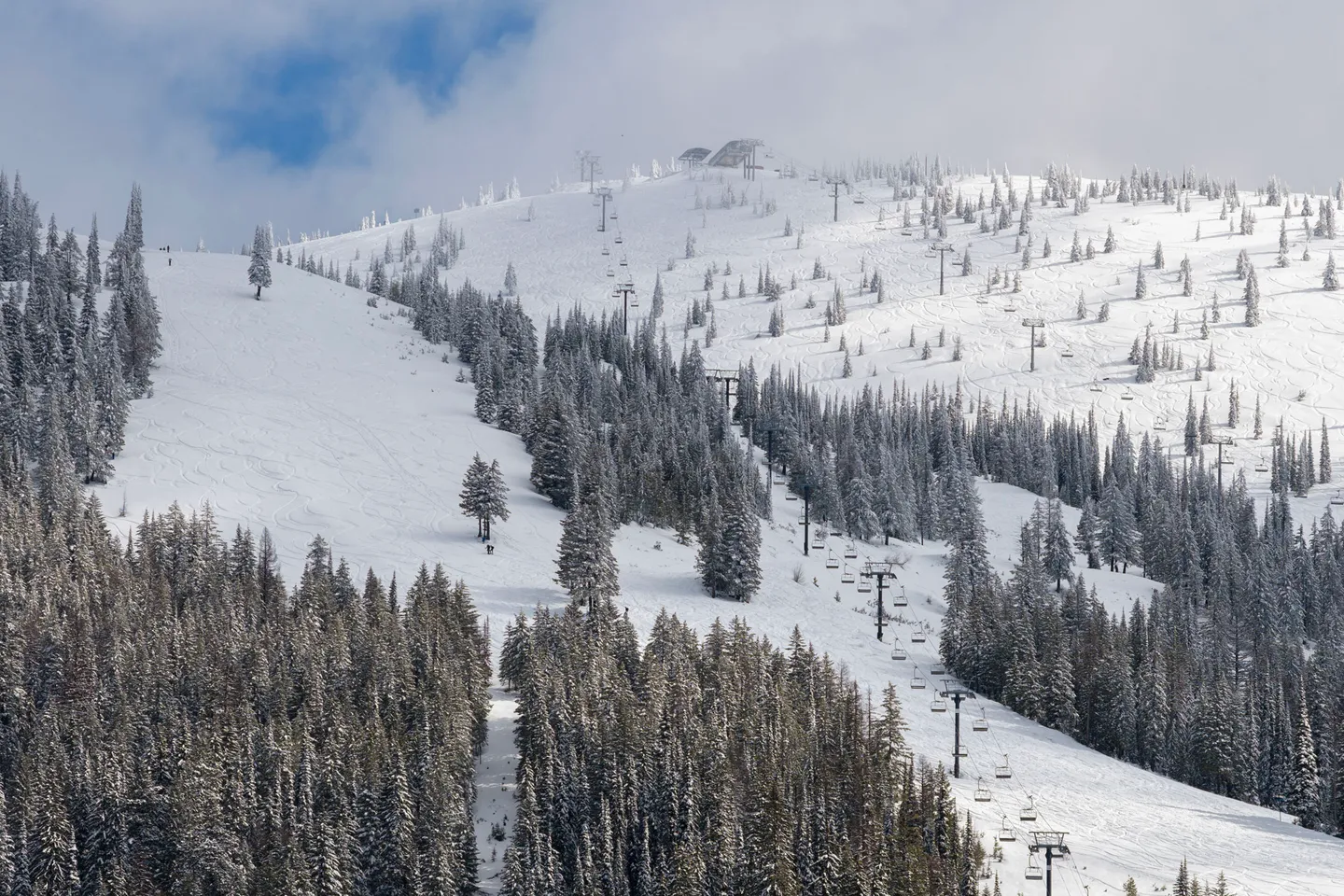 Snow-covered mountain ski slopes lined with pine trees and chairlift towers under a partly cloudy sky.