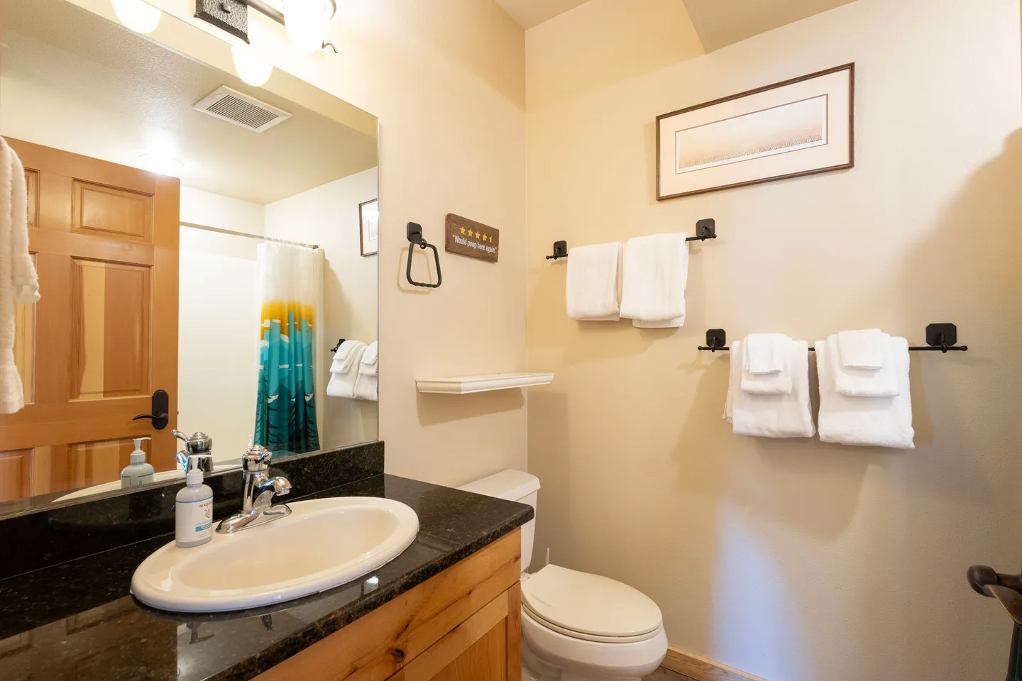 Bathroom with a vanity sink, toilet, towel racks, and a shower with a colorful curtain visible in the mirror.