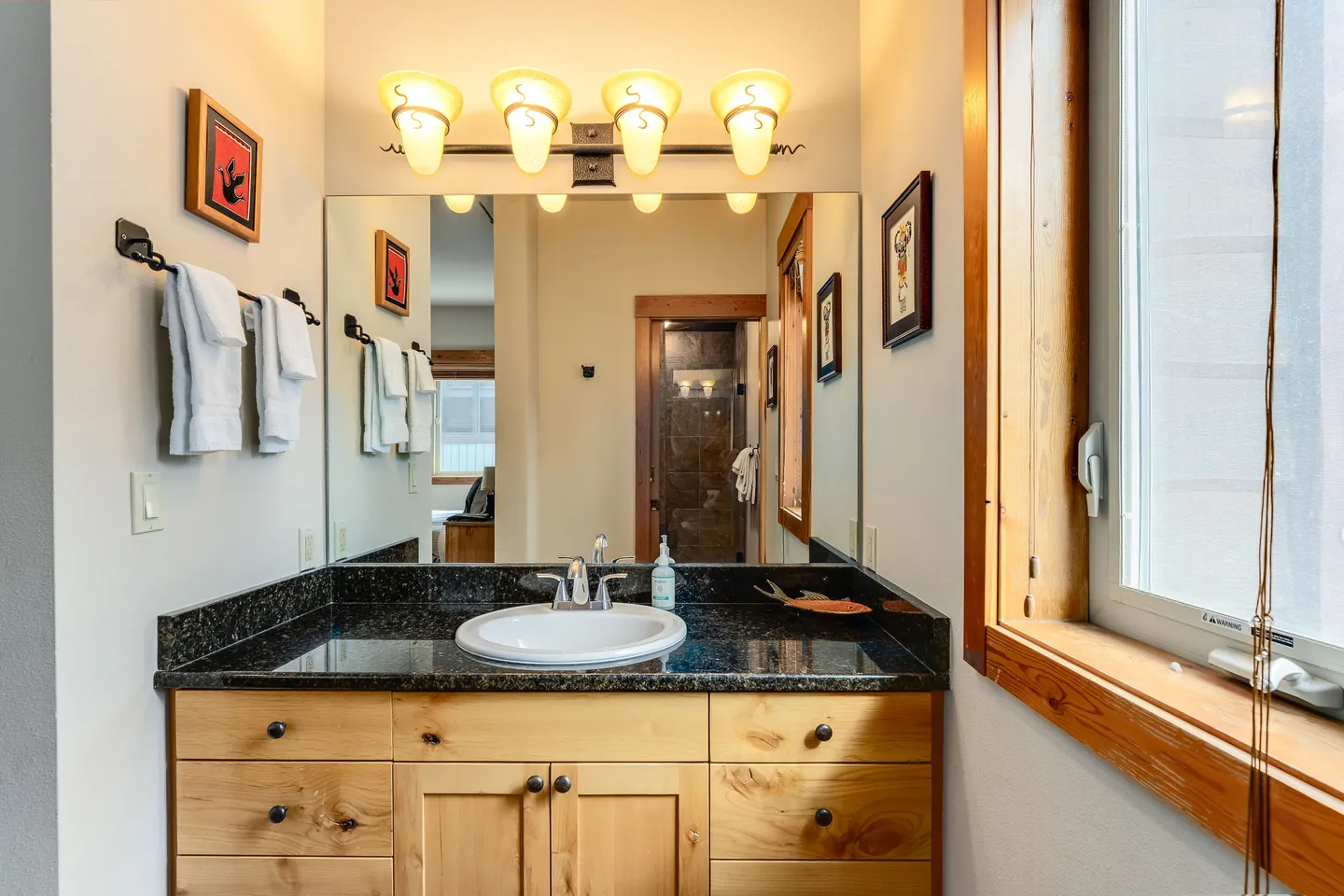 Bathroom vanity with a dark countertop, white oval sink, wood cabinetry, large mirror, and four-bulb overhead vanity lighting.