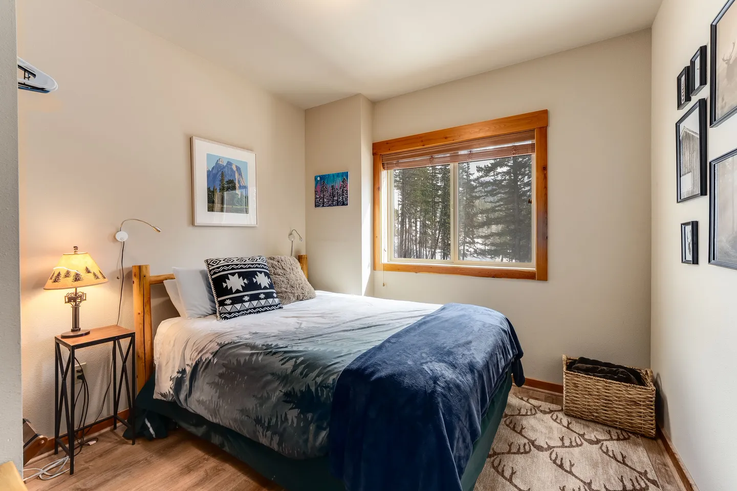 Bedroom with a wooden frame bed, rustic nightstand with a lamp, wood-trimmed window overlooking trees, and framed nature art on the wall.