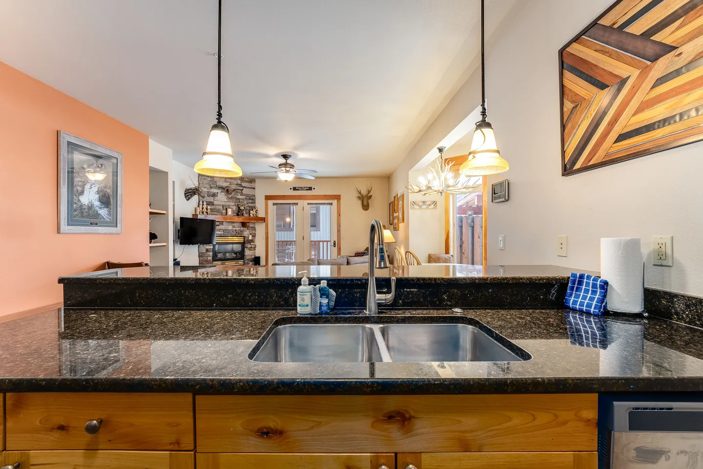 A kitchen counter with a double sink looks out into a living room with a stone fireplace.
