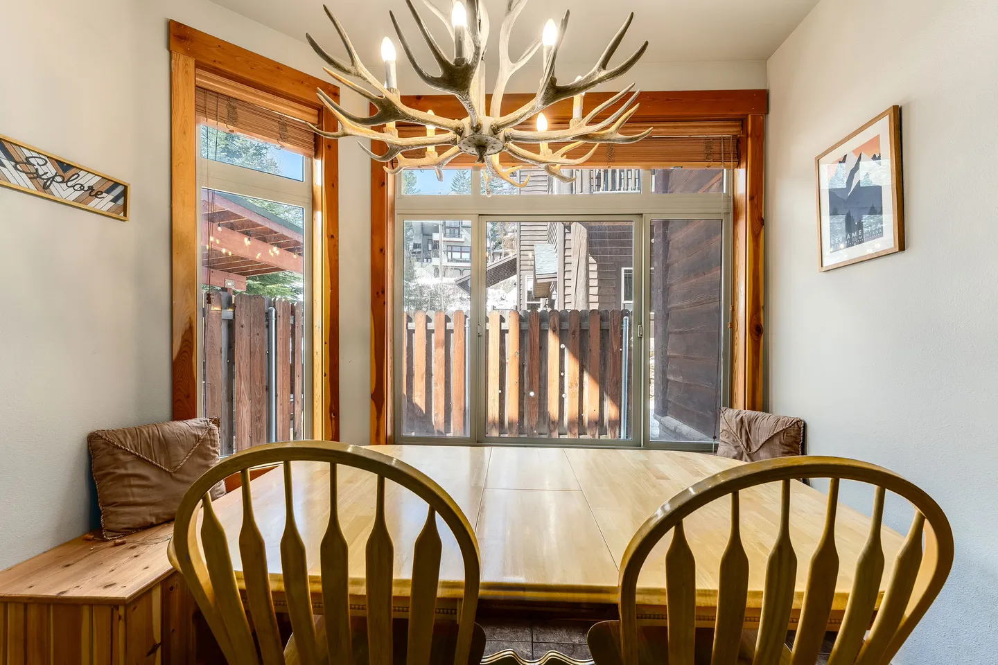 Dining area with a wooden table, spindle-back chairs, built-in corner benches, and an antler chandelier hanging below large windows.