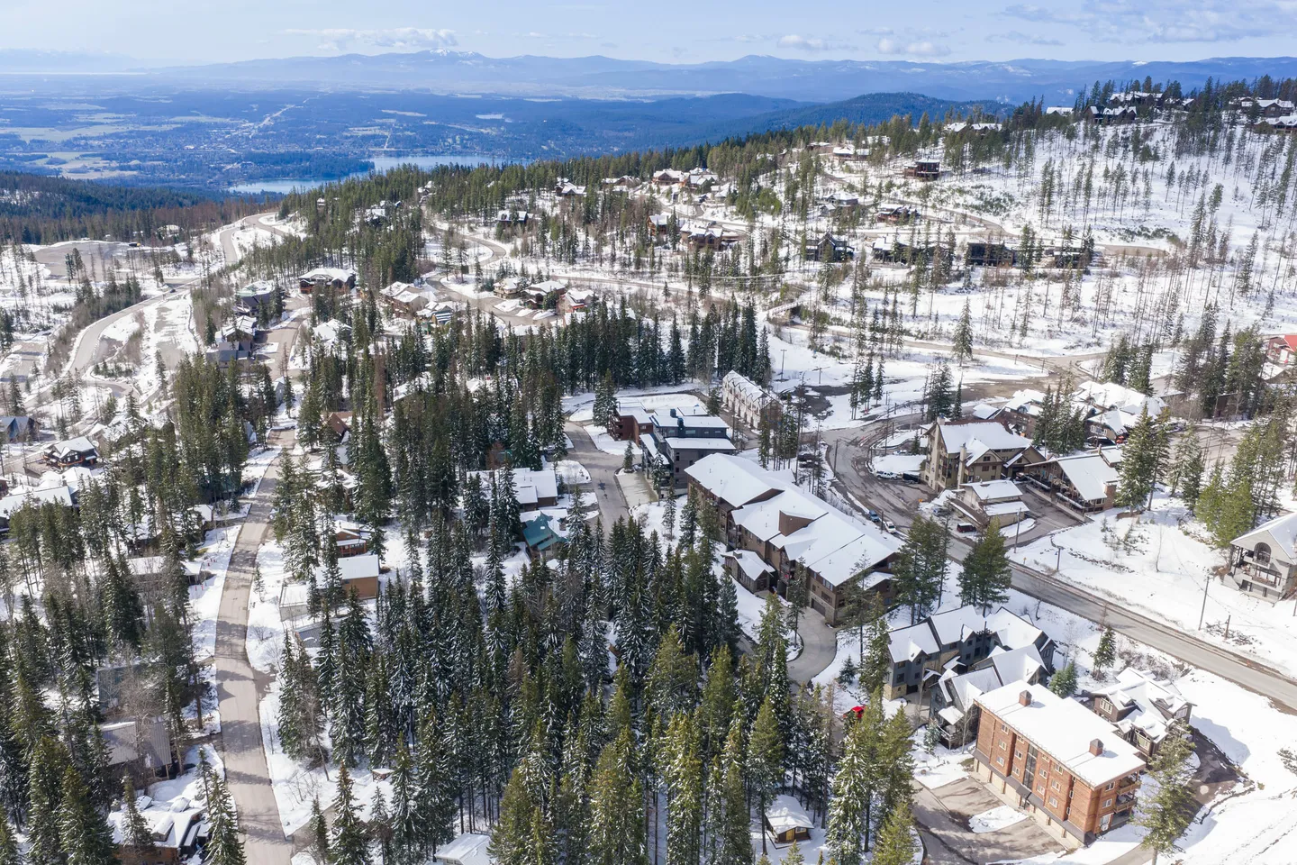 Aerial view of a snowy mountain village with roads, houses, and evergreen trees under a blue sky.