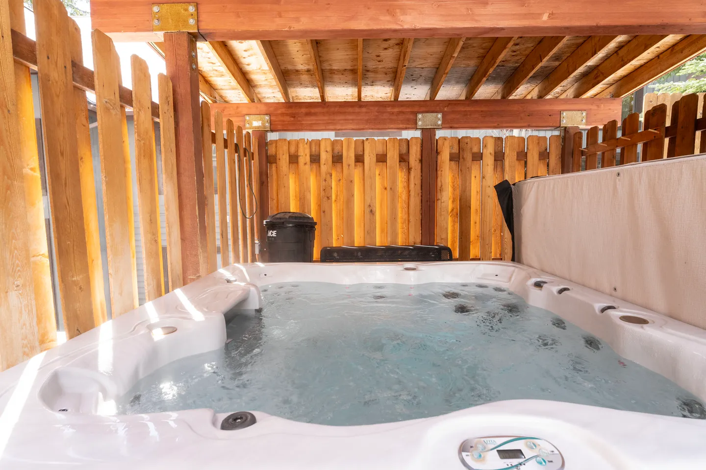 Outdoor hot tub filled with bubbling water under a covered wooden structure and enclosed by wood privacy fencing.
