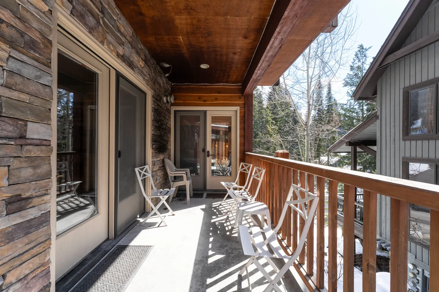 A covered outdoor balcony with stone veneer siding, wooden railings, and several white folding chairs overlooking trees.