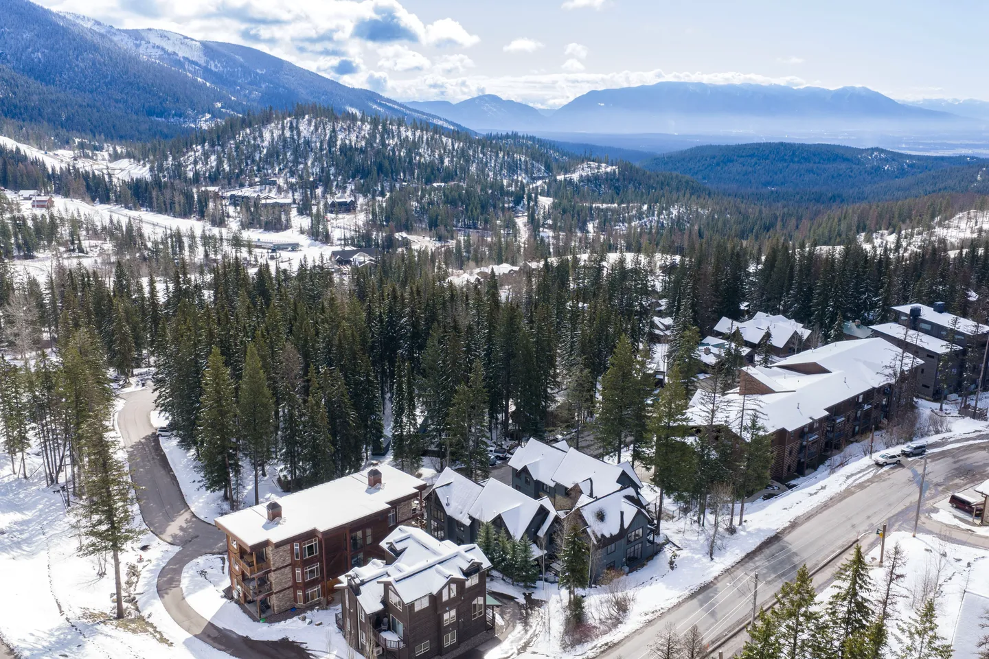 Aerial view of snow-covered mountain lodges surrounded by evergreen trees under a blue sky.