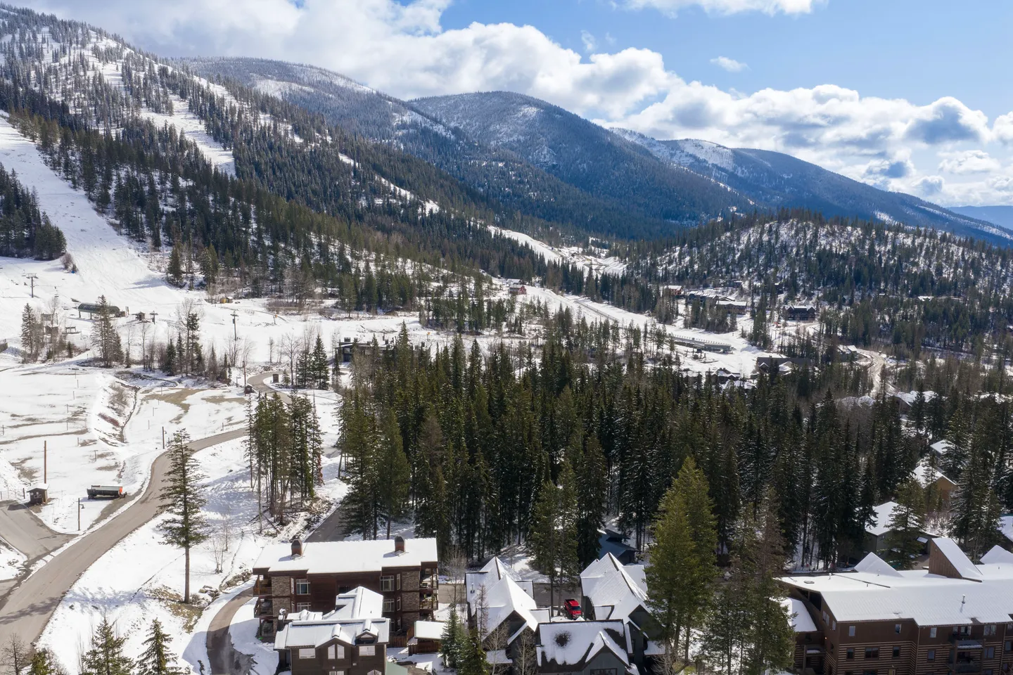 Aerial view of snow-covered mountain terrain, ski slopes, evergreen trees, and nearby resort lodges under a partly cloudy sky.