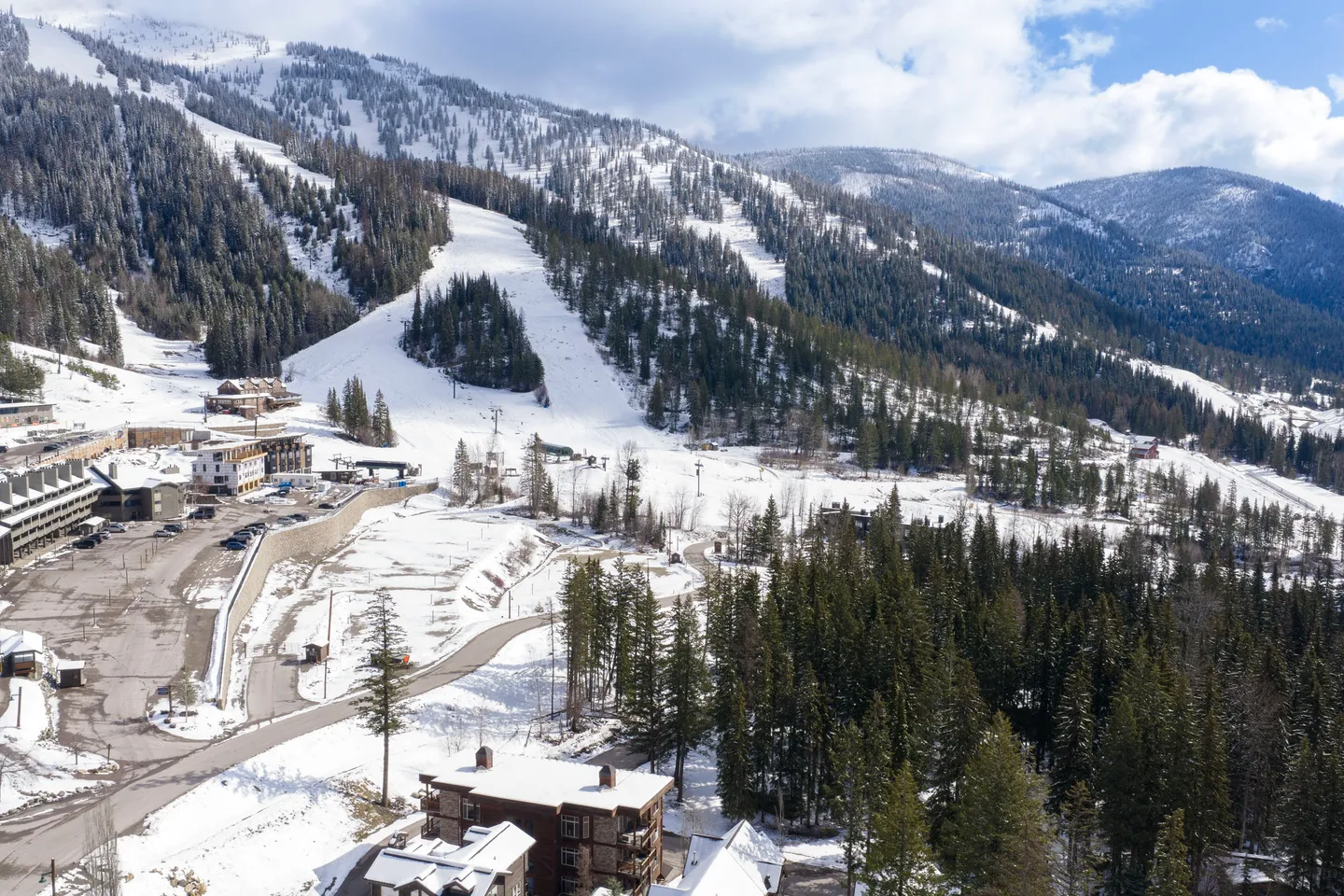 Aerial view of a snowy ski resort base area with ski runs carved into evergreen-covered mountains under a blue sky.