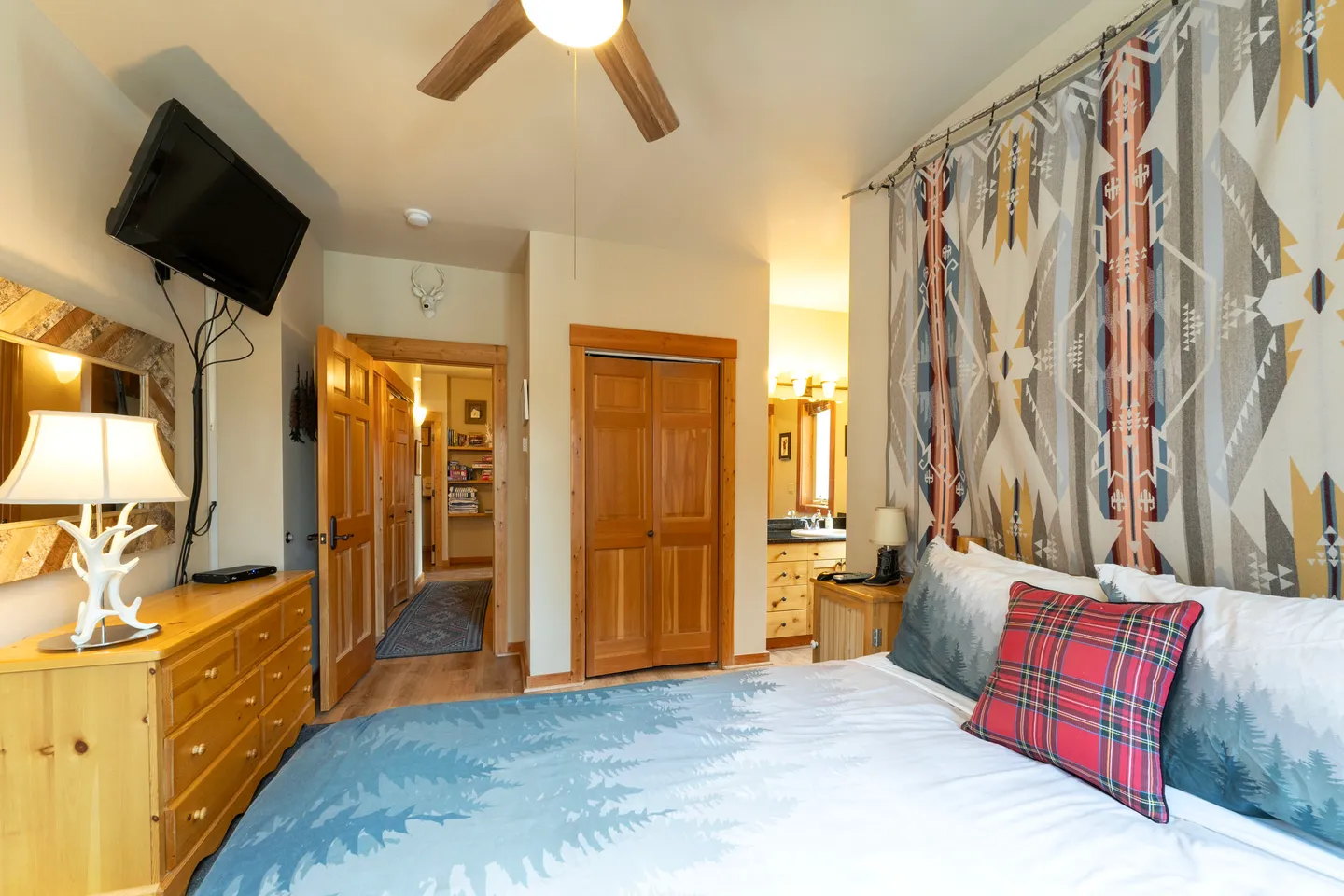 Bedroom featuring a bed with a southwestern-patterned blanket on the wall, wood dresser, wall-mounted TV, and an open doorway leading to a hallway and en suite bathroom.