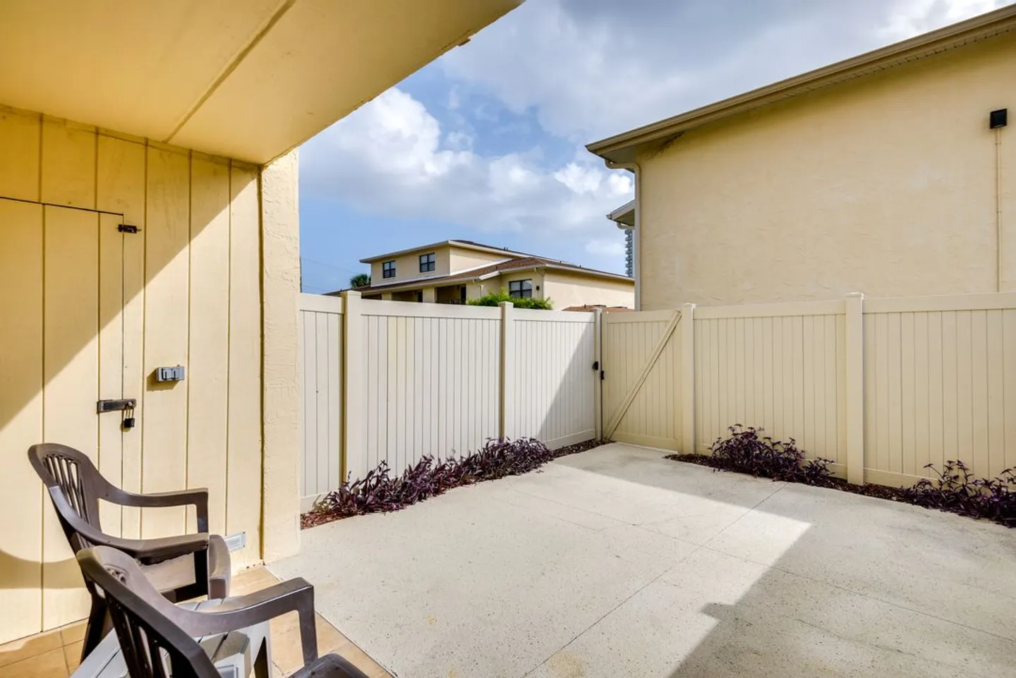 Fenced concrete patio with outdoor armchairs, a gate, and low purple plants along the fence line.
