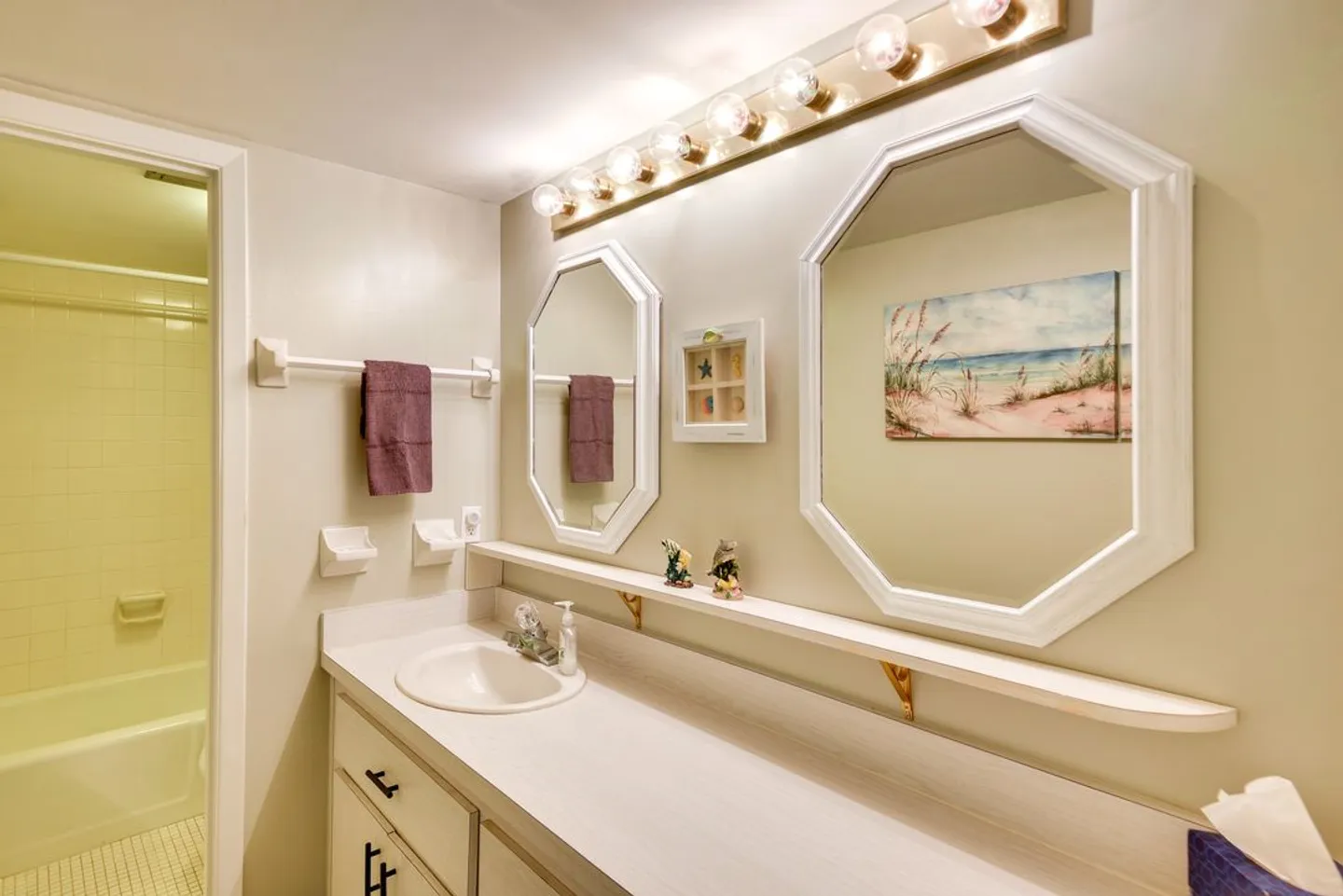 Bathroom vanity with a white countertop, two octagonal mirrors, a Hollywood-style light bar, and a glimpse into a tub-shower area.