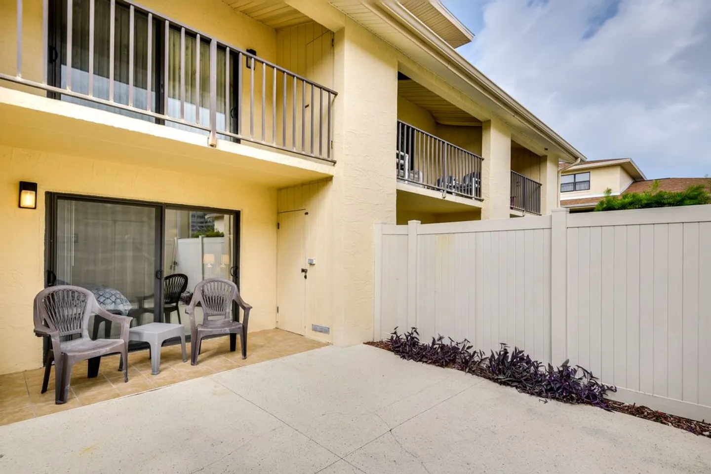 Enclosed outdoor patio with concrete paving, plastic armchairs, sliding glass doors, and a white privacy fence.