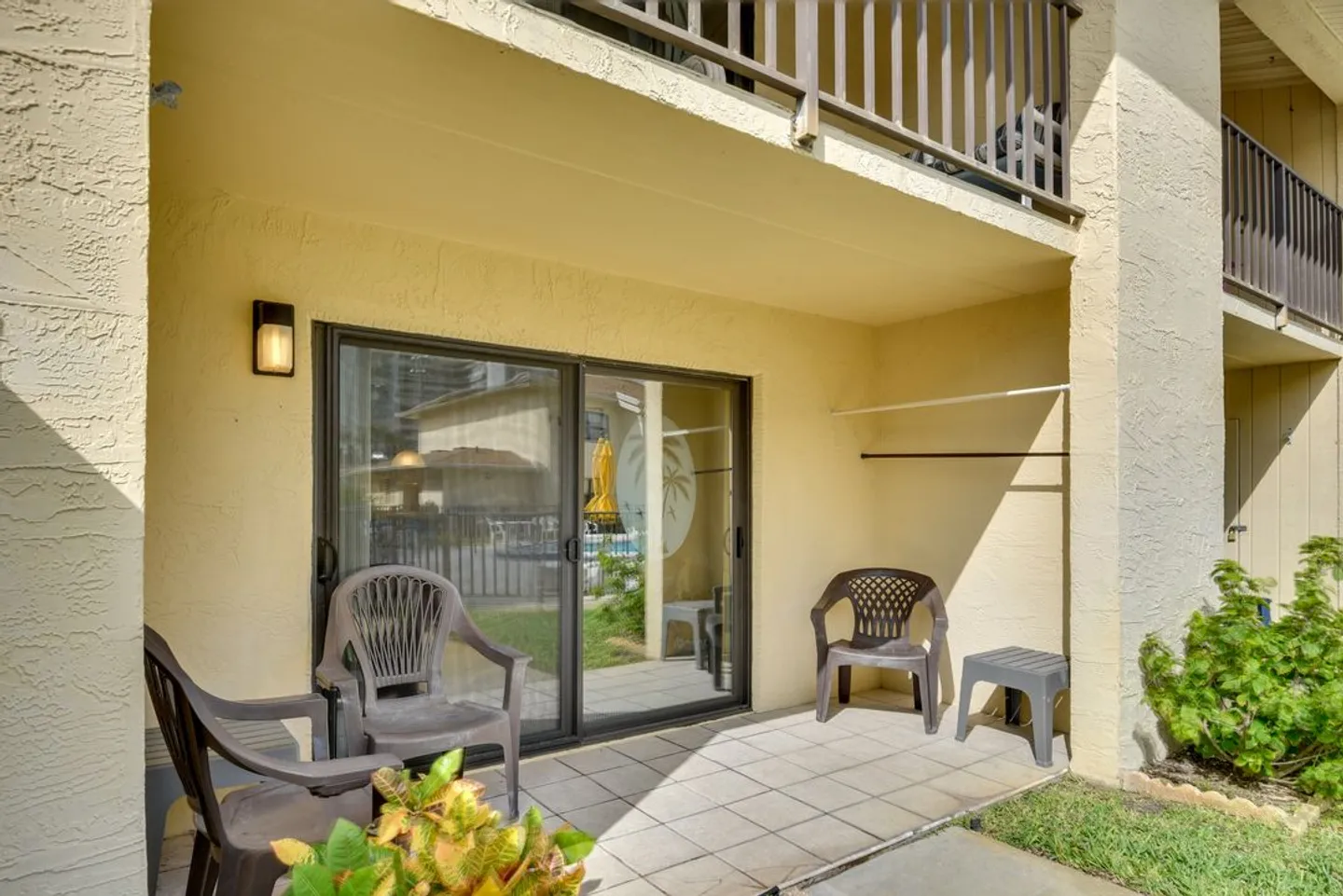 Ground-floor covered patio with tiled flooring, plastic chairs, and sliding glass doors under an upper balcony.