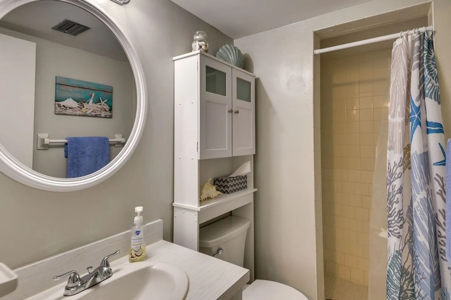 Bathroom showing a vanity with a circular mirror, an over-the-toilet storage unit, and a tiled shower stall with a patterned curtain.