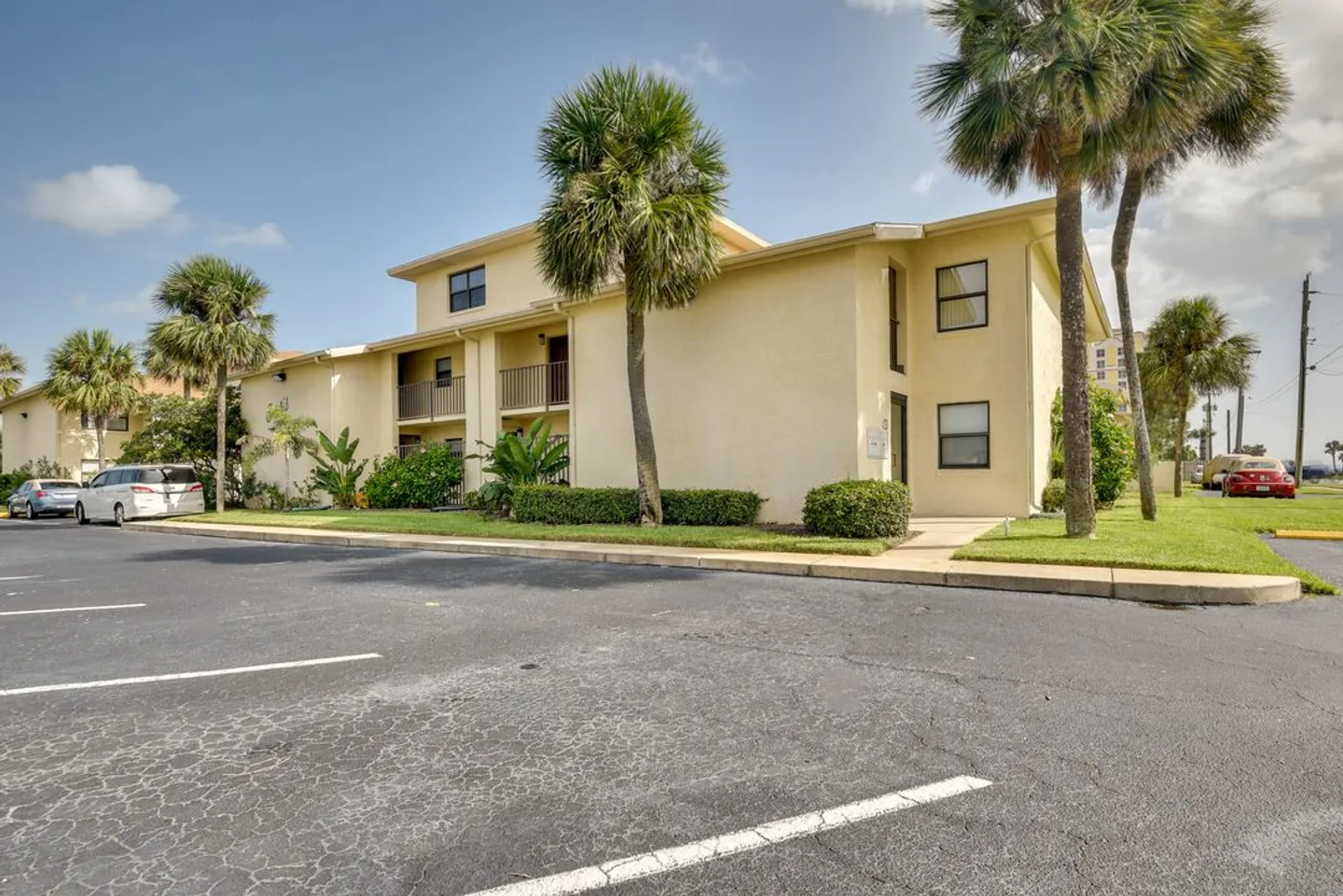 Exterior view of a two-story light yellow apartment building with palm trees, manicured hedges, and an asphalt parking lot.