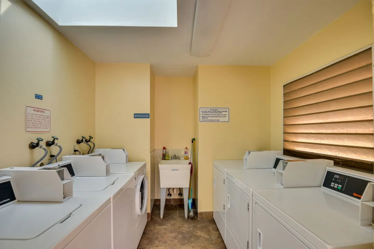 A shared laundry room with rows of commercial washing machines and dryers, a utility sink, and a window with horizontal blinds.