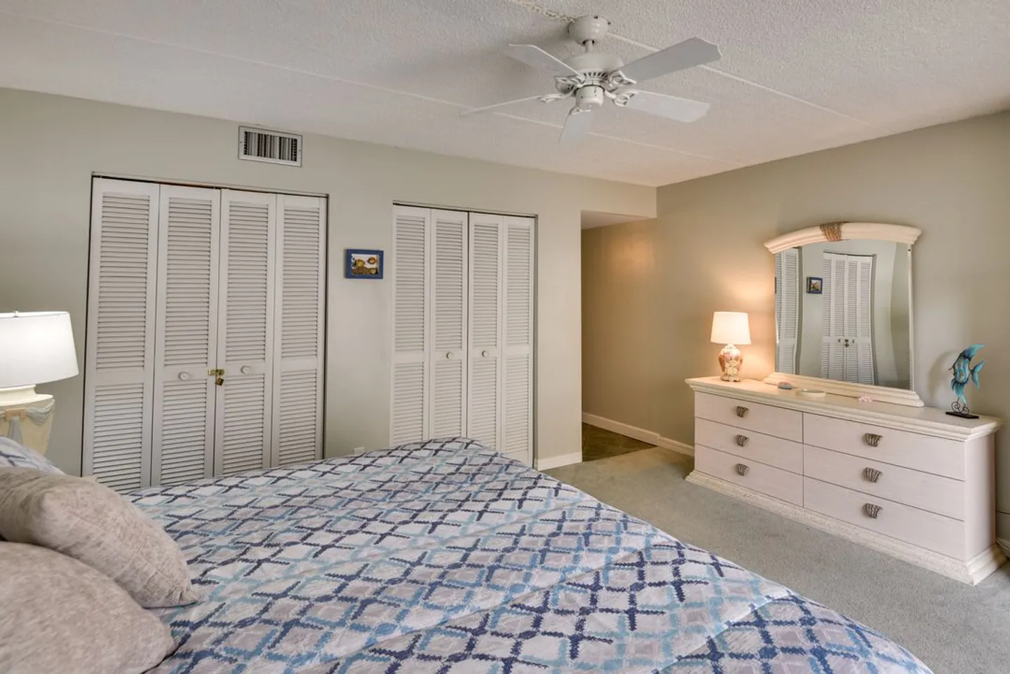 Bedroom featuring a made bed with blue-patterned bedding, white bifold closet doors, a ceiling fan, and a light-toned dresser with an attached mirror.