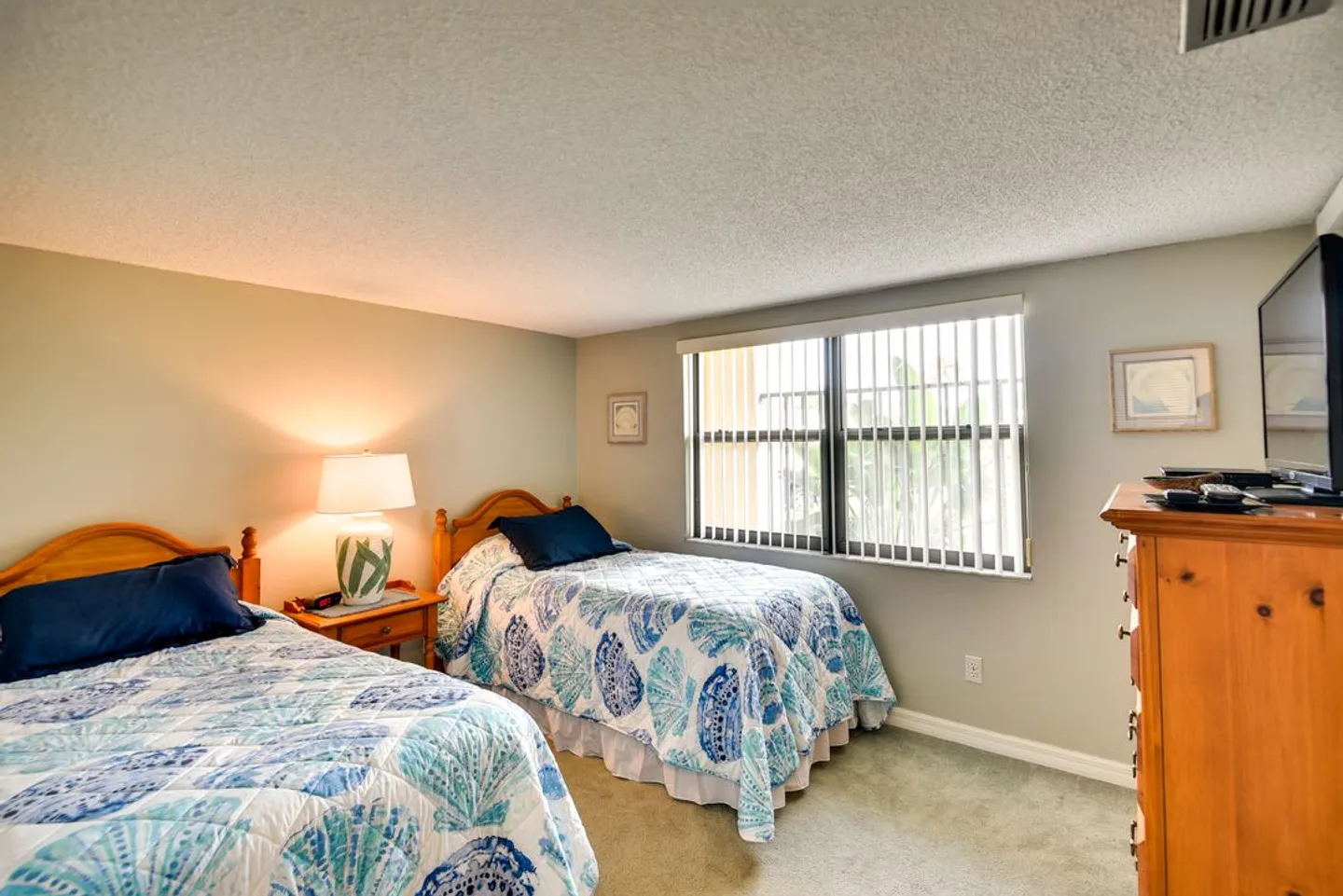 Bedroom with two twin beds dressed in blue and white seashell-patterned quilts, a nightstand with a lamp between them, and a window with vertical blinds.