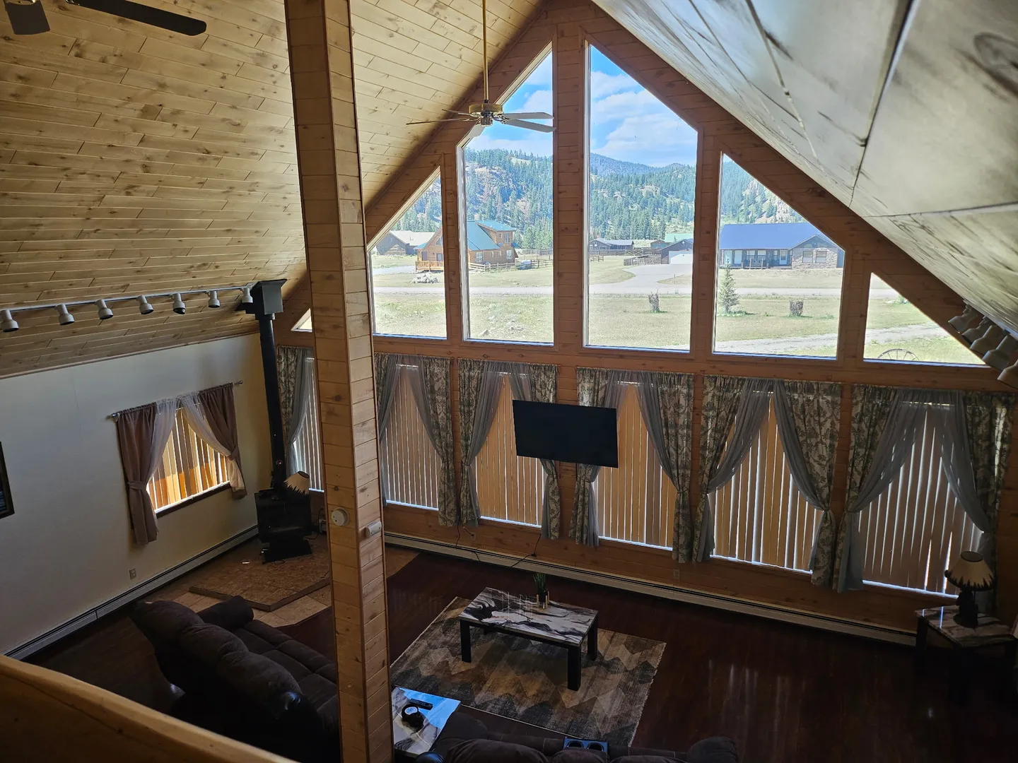 High-angle view of a living room with vaulted wood ceilings, a large wall of windows, a wood stove, and a seating area.