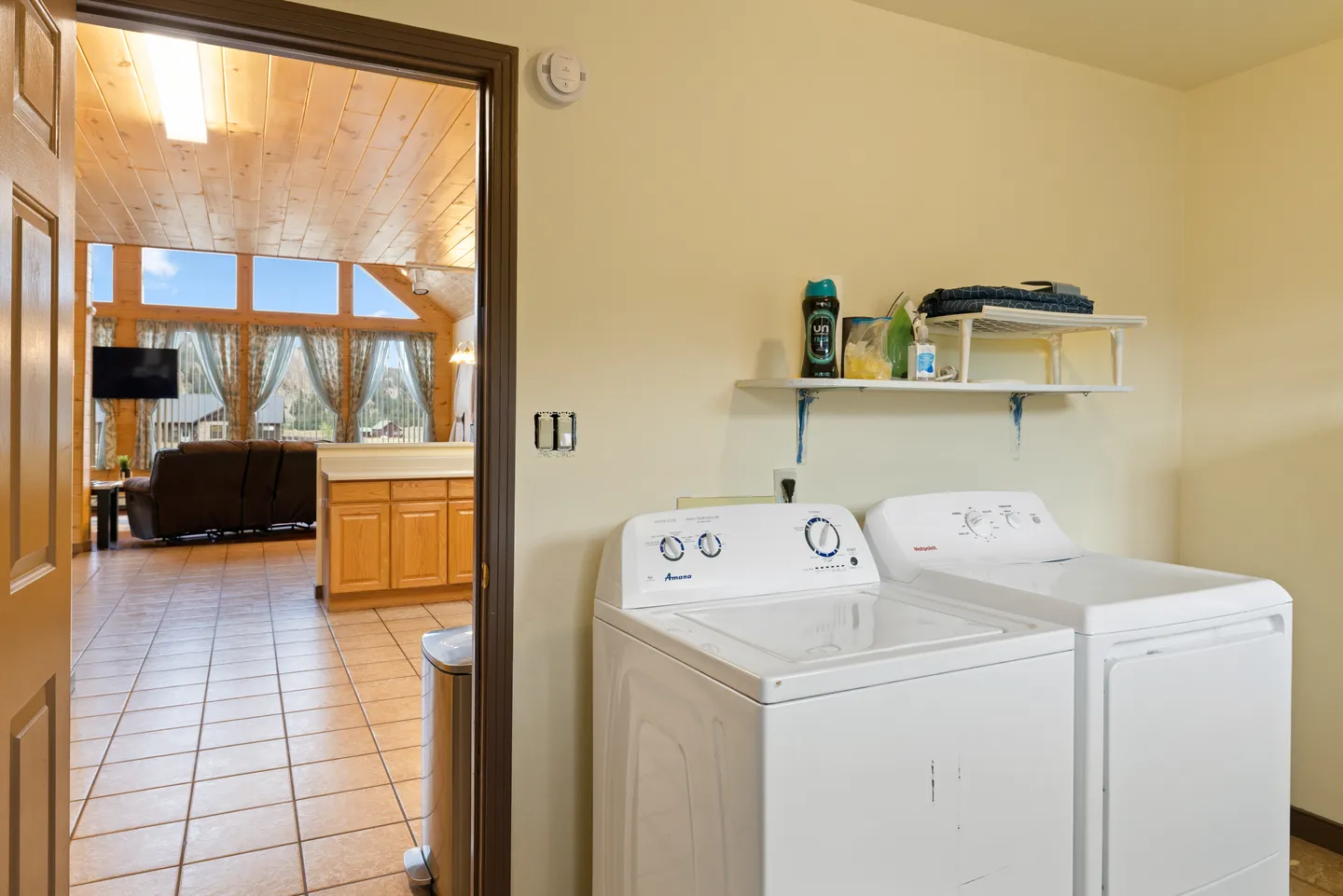 A laundry room with a white washer and dryer, looking out into a tiled open-concept living and dining area with wood ceilings.