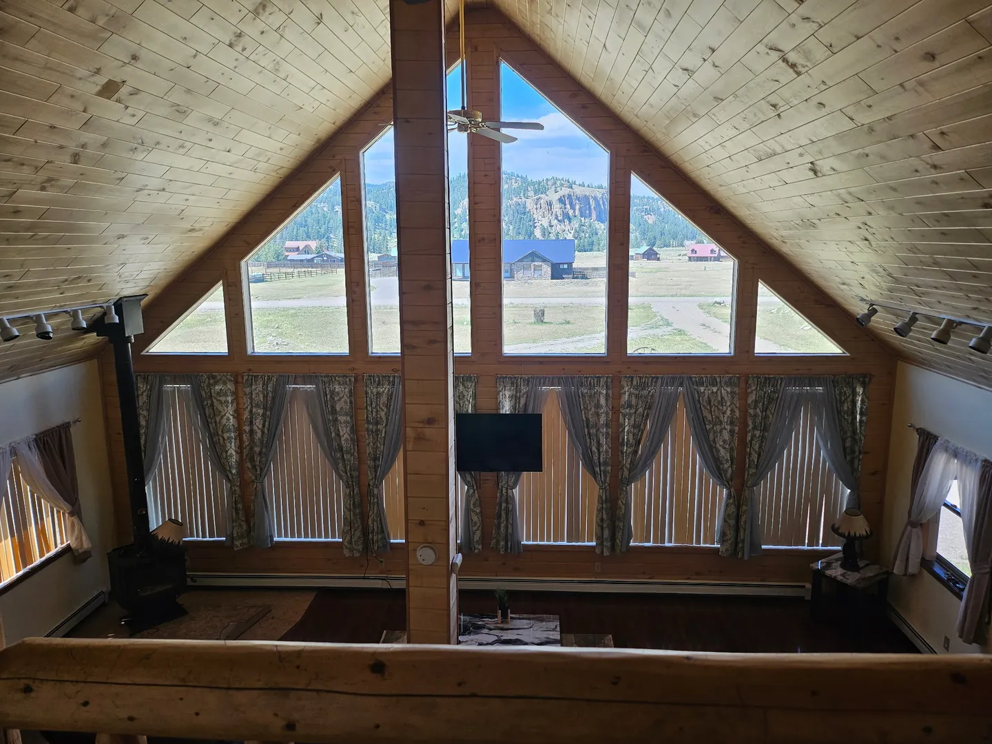 A high-angle view from a loft looking down into a living room with vaulted wood ceilings and a large A-frame wall of windows.