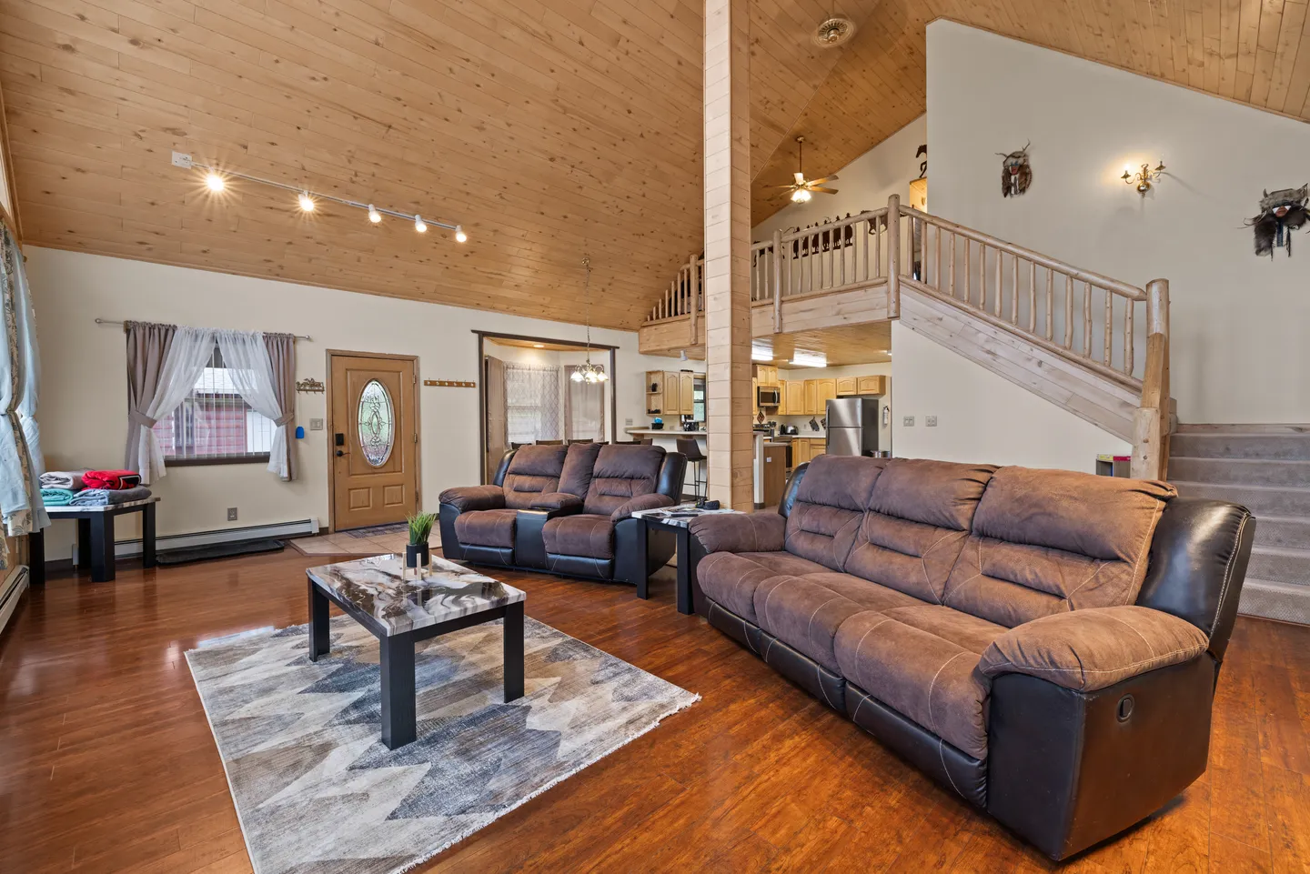 Open living area with wood-paneled vaulted ceiling, brown plush sofas, coffee table, wood-look flooring, and a staircase leading to an upper loft.