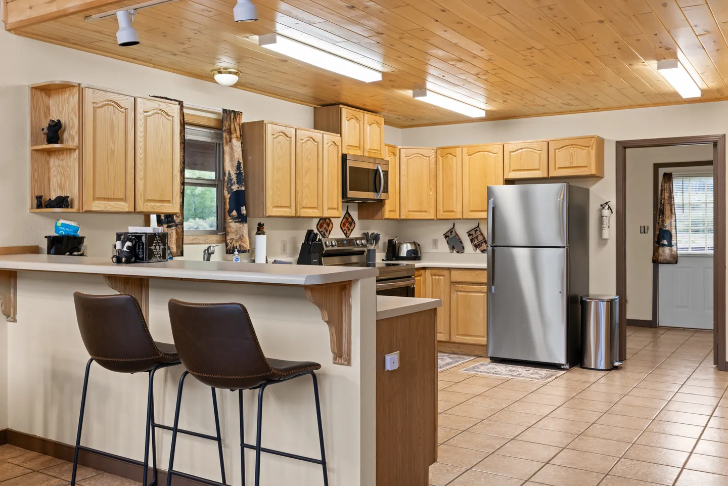 Kitchen with wooden cabinetry, a wood-paneled ceiling, tiled floor, a peninsula counter with barstools, and stainless steel appliances.