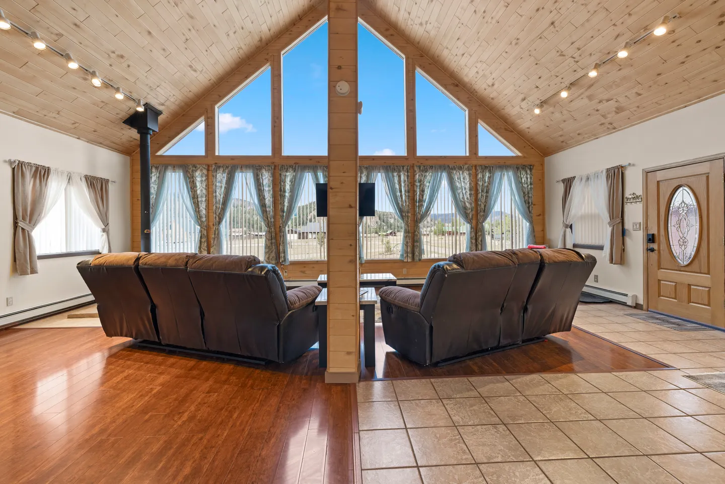Living room with vaulted wood-paneled ceiling, large A-frame windows, and two brown reclining sofas facing the outdoors.