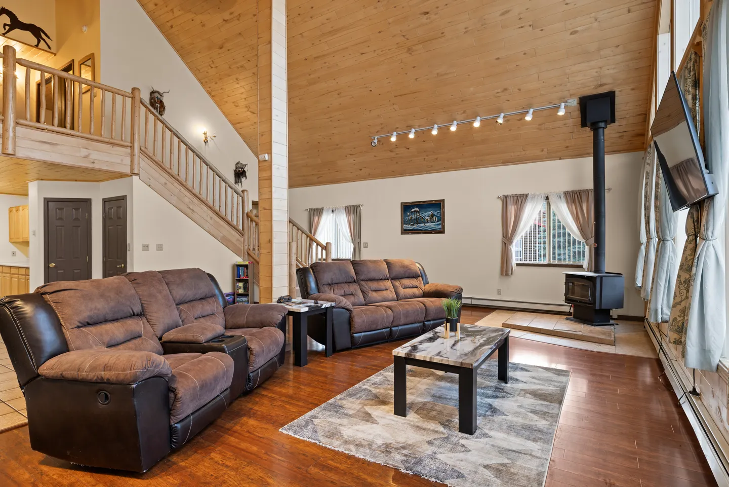 Living room with high wood-paneled vaulted ceiling, brown reclining sofas, coffee table, area rug, wood stove, and a wooden staircase leading to an upper loft.