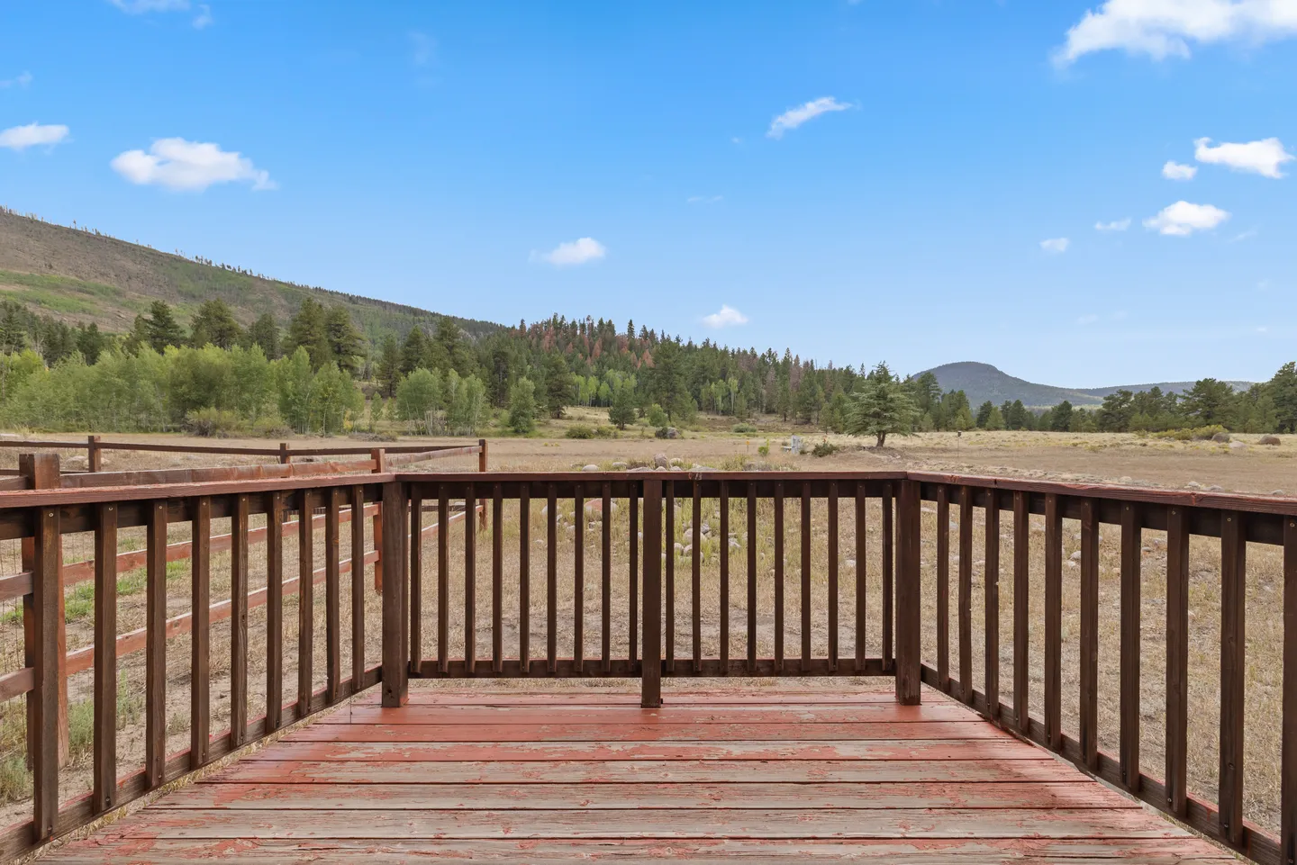 A wooden outdoor deck with brown railings overlooking a wide meadow and tree-covered hills under a blue sky.