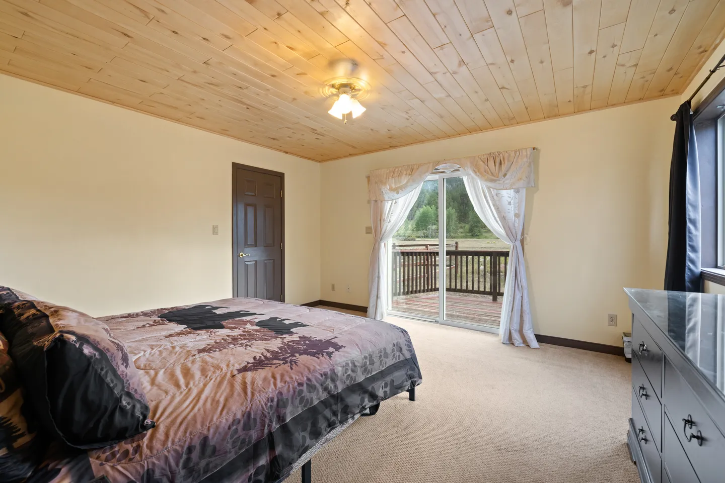 Bedroom featuring a wood-plank ceiling, carpeted floor, bed with wilderness-patterned bedding, and sliding glass doors to a deck.