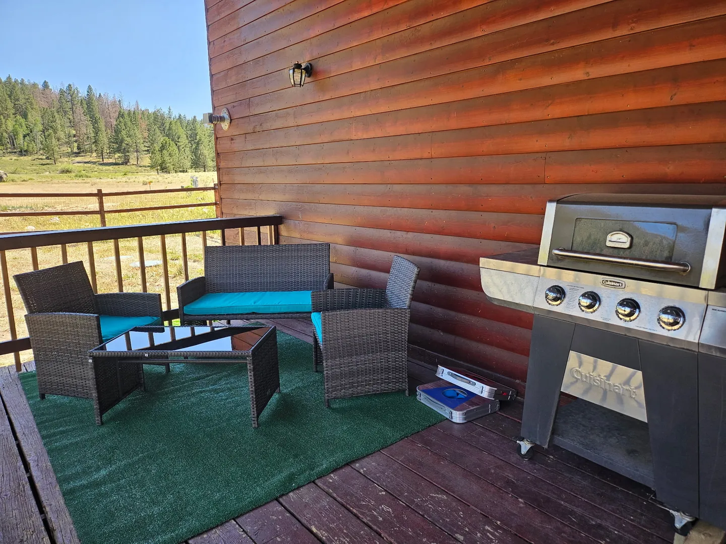 Outdoor wooden deck featuring a wicker seating set with turquoise cushions, a glass-top coffee table, and a stainless steel grill against log siding.