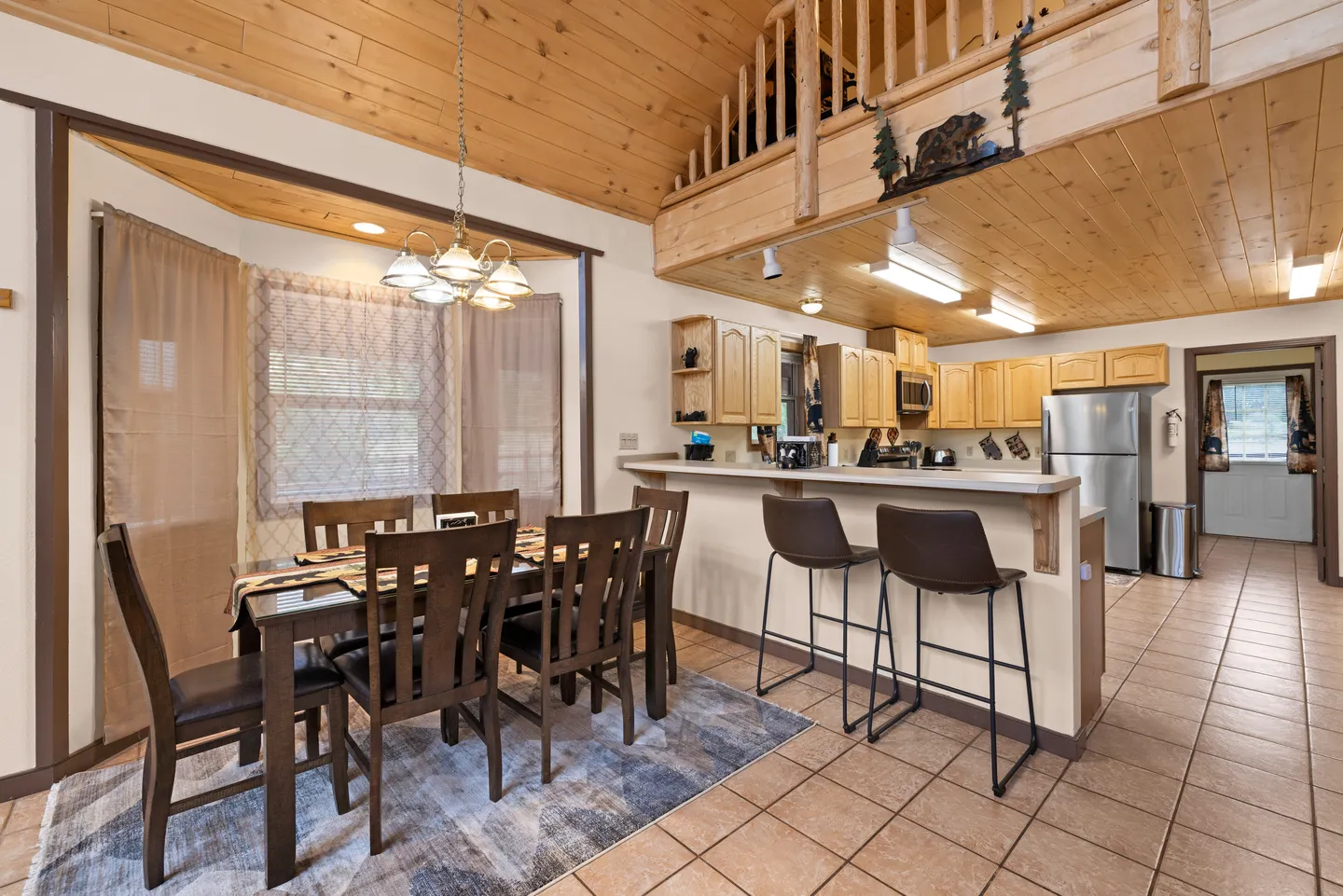 Dining area with a wooden table and chairs adjacent to a kitchen with a breakfast bar, wooden ceiling, and loft above.
