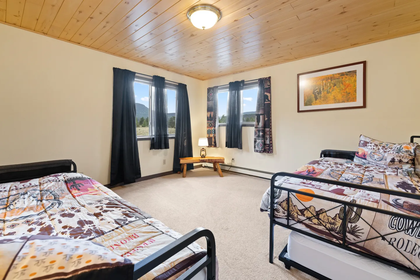 Bedroom with wood-paneled ceiling, carpeted floor, two twin beds with Western-themed bedding, and two windows looking out toward mountains.