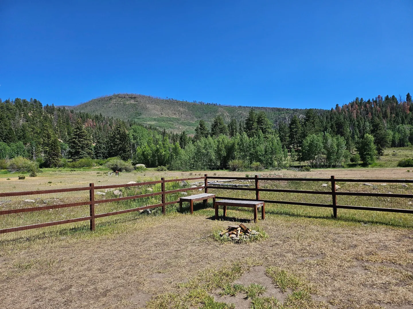 Outdoor fire pit area with wooden benches beside a fence overlooking a mountain meadow.
