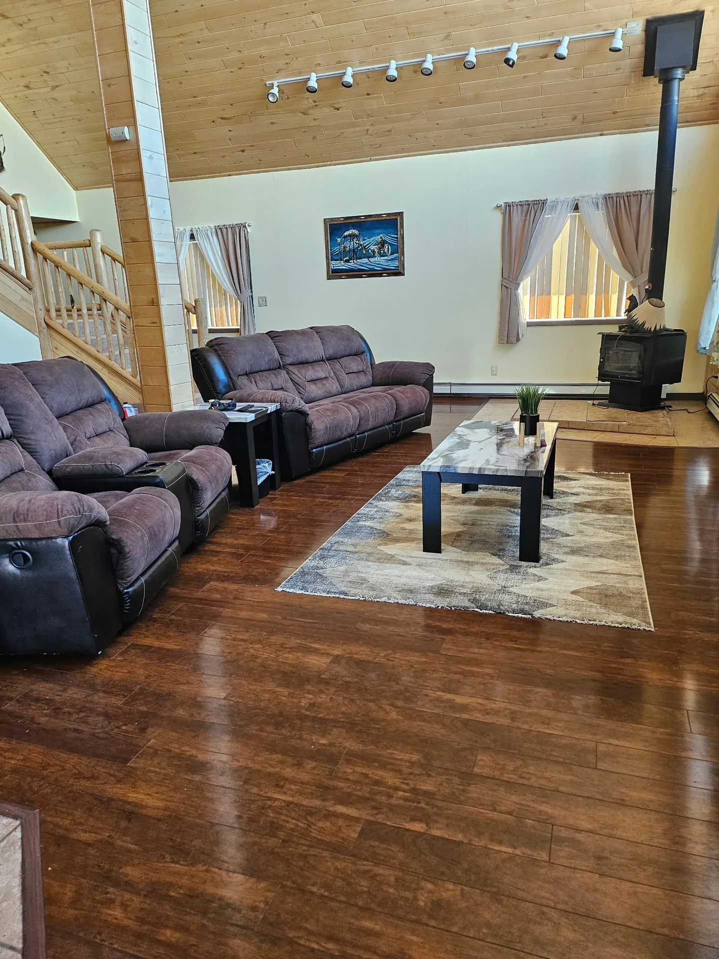 Living room with wood-paneled ceiling, dark reclining couches, a coffee table on a patterned rug, and a corner wood stove.