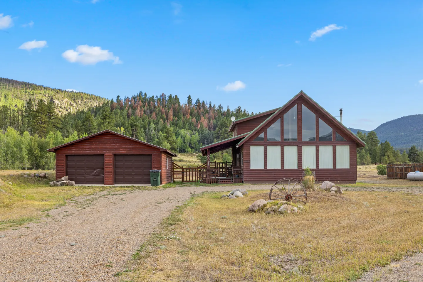 Exterior view of a log cabin with a two-car garage set against a wooded mountain backdrop under a blue sky.