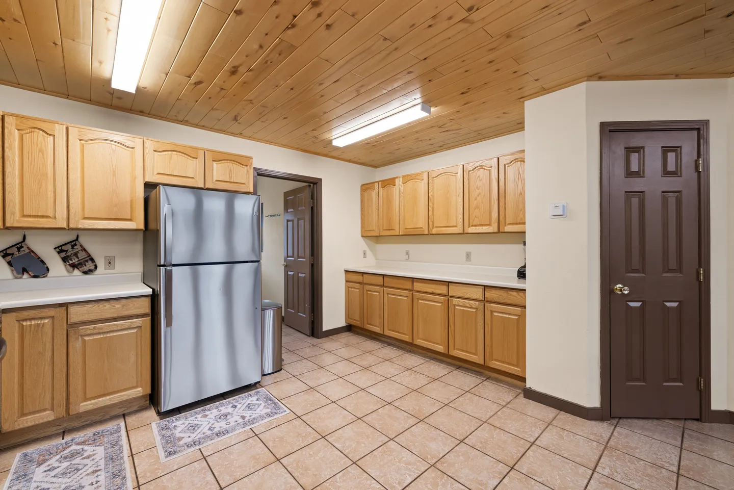 Kitchen area with light wood cabinets, stainless steel refrigerator, tile floor, and wood-paneled ceiling.