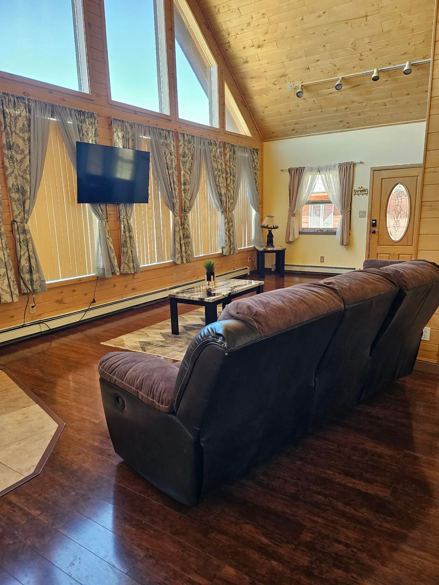 Living room with vaulted wood-paneled ceiling, tall windows with blinds and curtains, a wall-mounted TV, and a brown reclining sofa.