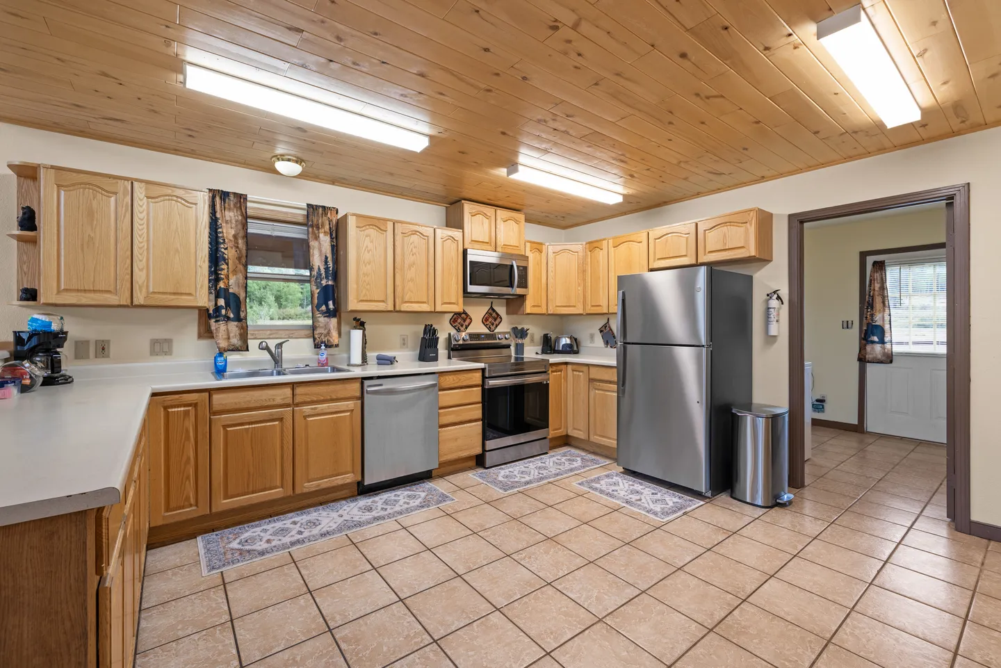 A kitchen with light wood-paneled ceiling, light oak cabinets, tiled floor, and stainless steel appliances including a refrigerator, stove, microwave, and dishwasher.