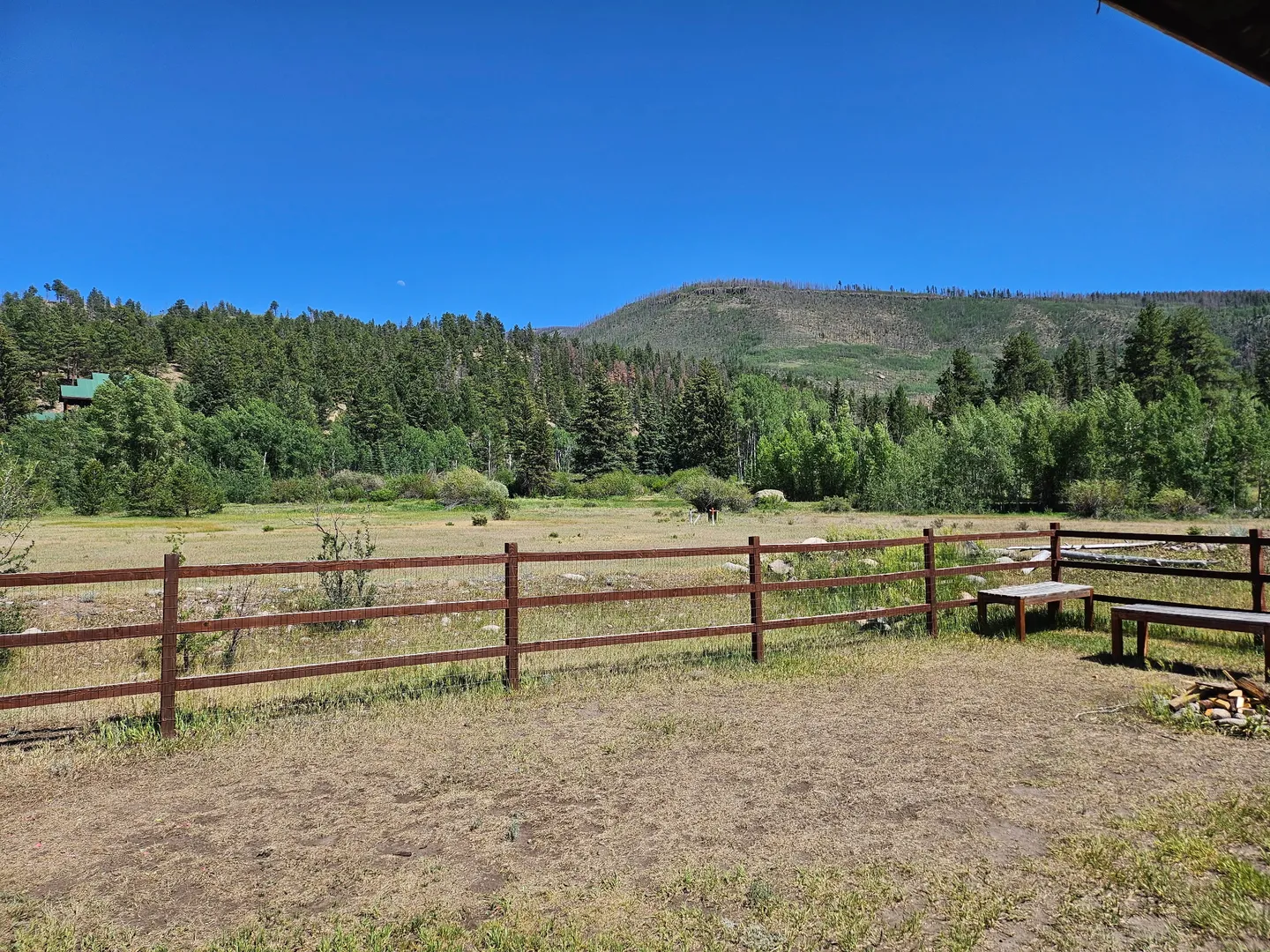 Open yard with a rustic wooden split-rail fence overlooking an open field, evergreen forest, and a mountain under a clear blue sky.