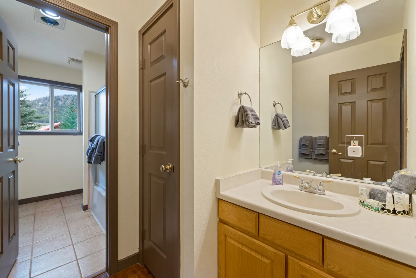 Bathroom with an oak vanity, large mirror, and an adjoining room with tiled floors and a window.